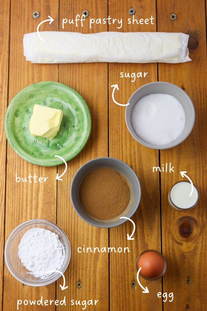 gathering ingredients for puff pastry cinnamon pinwheels: puff pastry sheets, sugar, powdered sugar, cinnamon, butter, an egg and milk 