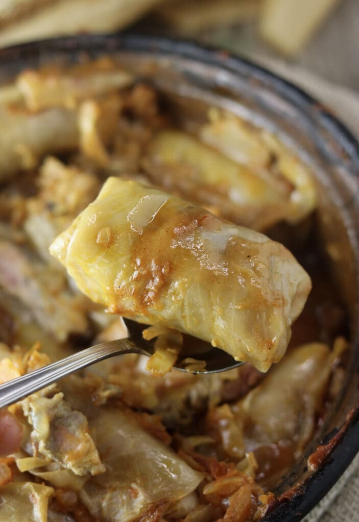A single cabbage roll with buckwheat on a large spoon with sauce and a whole baking pot with stuffed cabbage rolls in the background.