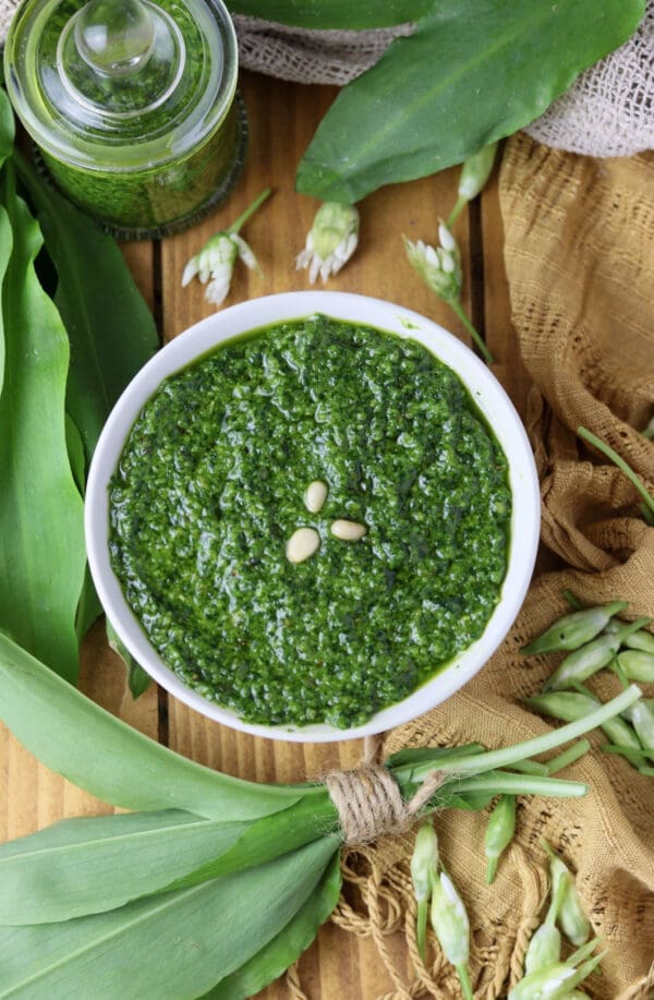 A bowl of pesto with wild garlic and toasted pine nuts with a glass jar and wild garlic leaves in the background.